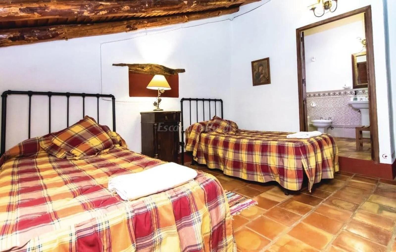 Una habitación con dos camas, techo de madera, una lámpara, un pequeño armario y un baño visible a través de una puerta abierta.