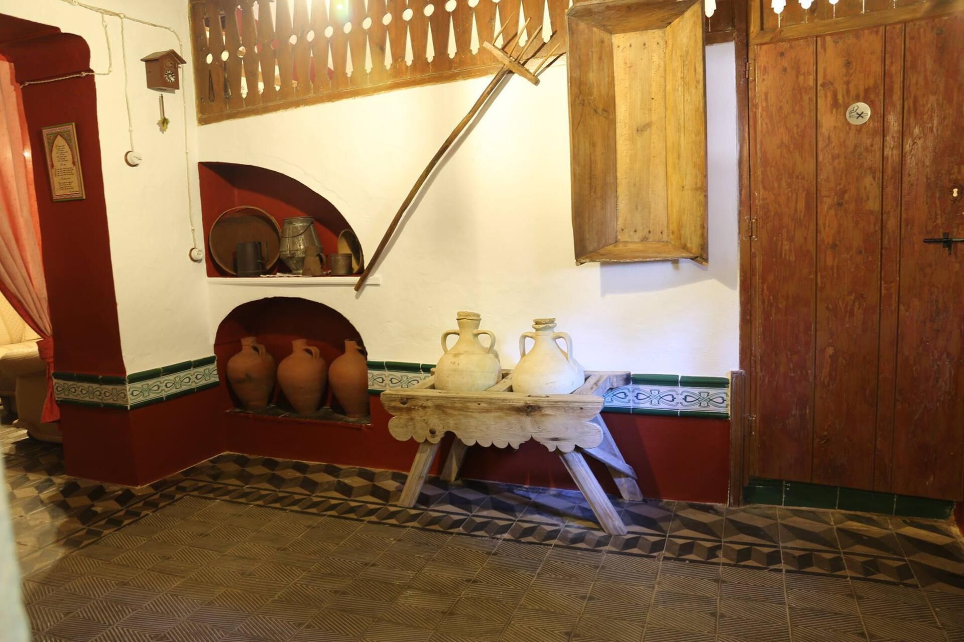 A room with a tiled floor, a wooden door, a shelf with pottery, and a wall-mounted clock.