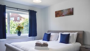 Een netjes opgemaakt bed met kussens, een nachtkastje, een raam met gordijnen, een radiator en een schilderij aan de muur.