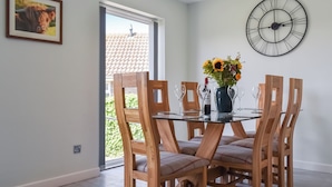 Een eetgedeelte met een glazen tafel, houten stoelen, een bloemenvaas en een wandklok.