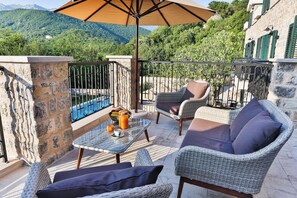 A balcony with wicker furniture, a table with a drink, and a view of green mountains. - Boutique hotel Ruzmarin Garden&SPA (Prijevor)