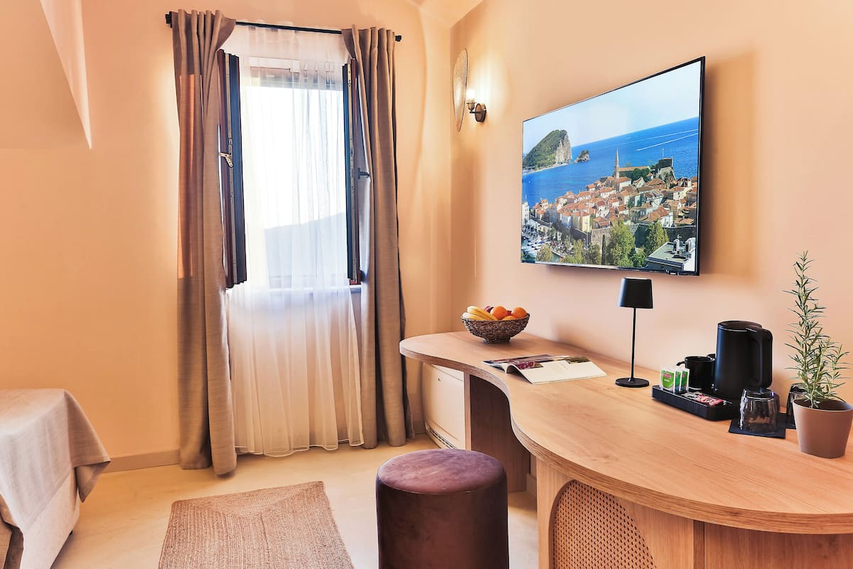 A hotel room with a wooden desk, a view of a coastal town, a window with curtains, and a potted plant.