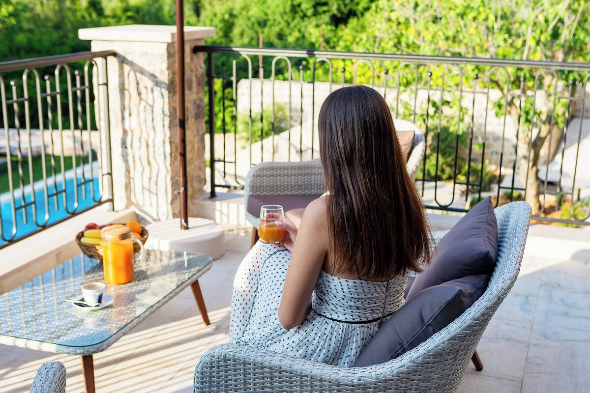 A woman sitting on a wicker chair with cushions, holding a glass of orange juice, on a balcony with a pool view.