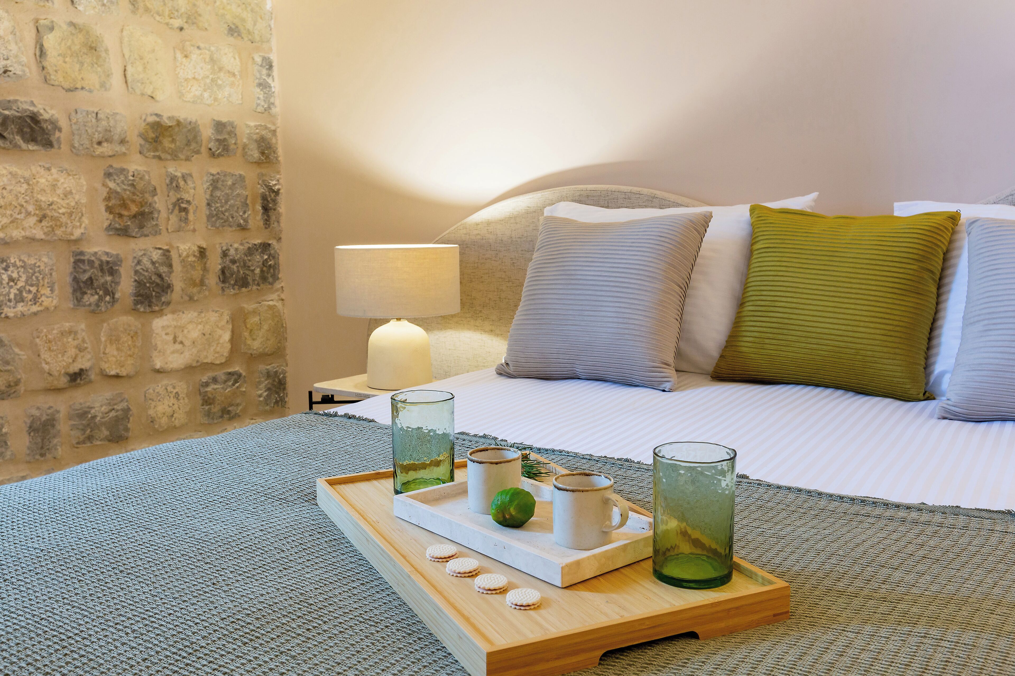 Una cama bien hecha con almohadas, una mesita de noche con una lámpara y una bandeja con vasos y una lima.