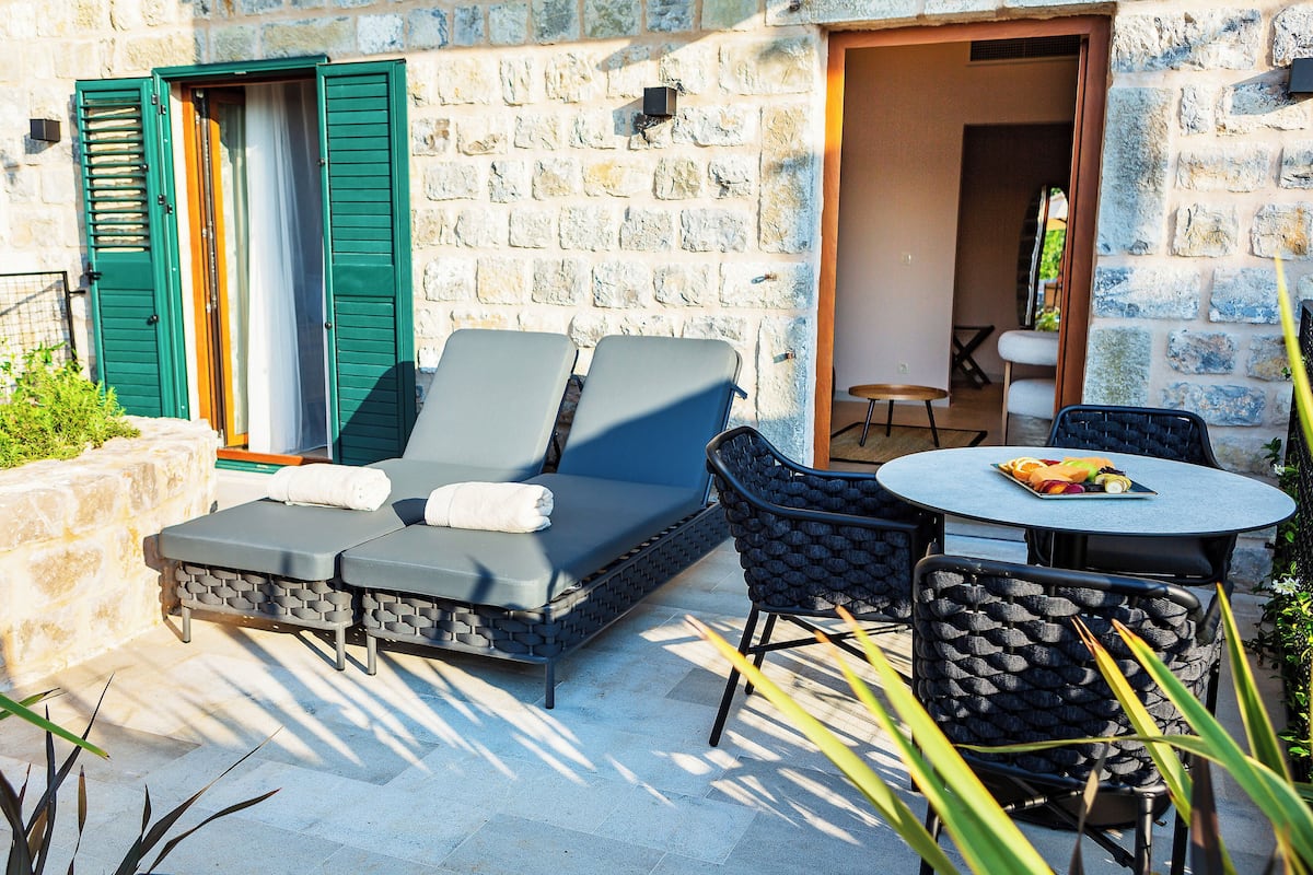 A stone-walled outdoor seating area with a sofa and chairs, a table with fruit, and a door with green shutters.