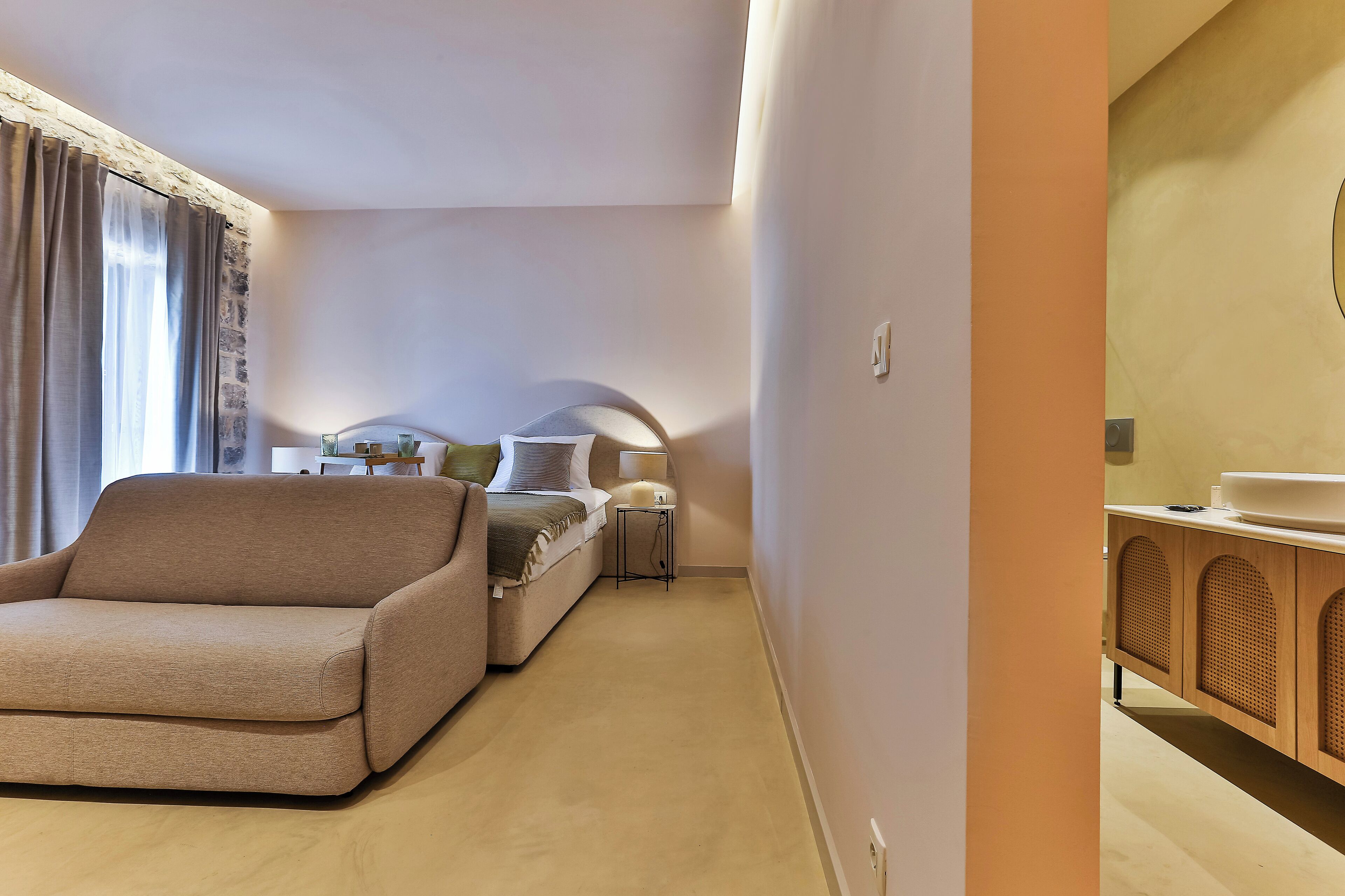 Una habitación de hotel moderna con sofá, una mesita de noche pequeña, lámpara y un mueble de madera.