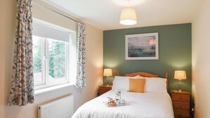 A bedroom with a bed, two bedside lamps, a window with curtains, a radiator, and a painting on the wall. - Tranquil lodge with sea views. (Plas Panteidal, near Aberdovey)