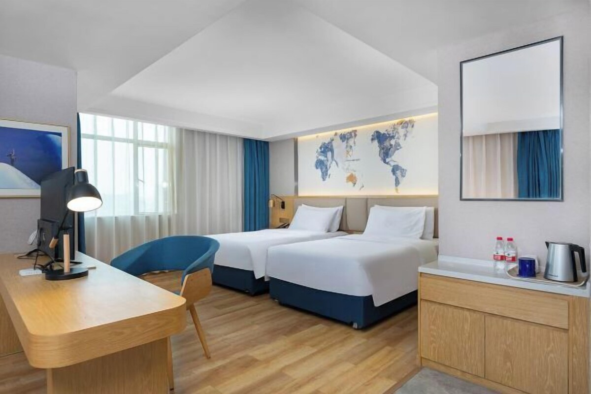 Habitación de hotel con dos camas, un escritorio con lámpara, una silla, un mapa mundial en la pared, un espejo y una kitchenette con hervidor y tazas.