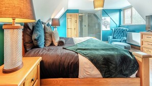 Un dormitorio con una cama, dos lámparas de noche, una mesita de noche de madera, un sillón azul, un aparador y un tragaluz.