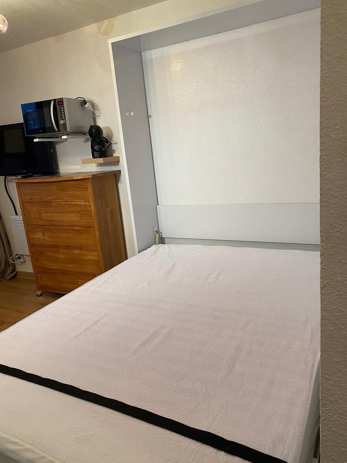 Ein Schlafzimmer mit einem Bett, einer Holzkommode, einer Mikrowelle und einem kleinen Tisch mit Wasserkocher und Kopfhörern.