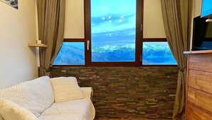 A room with a wooden floor, a white sofa, a stone wall, and a window with a view of snow-capped mountains. - Studio Corbier Zodiaque Pool and Ski at the foot of the slopes  (Villarembert)