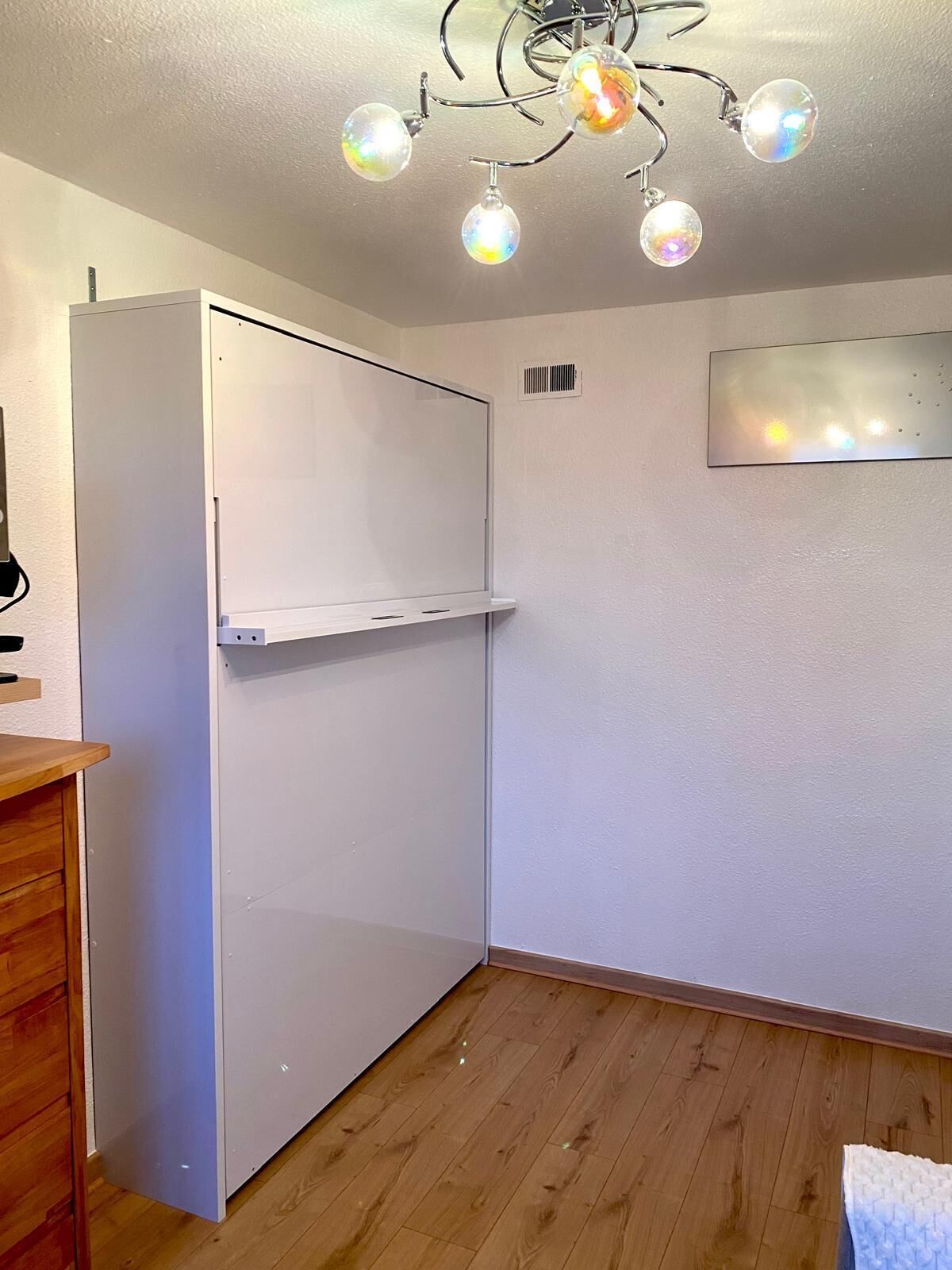 A room with a white wardrobe, a wooden floor, a white ceiling with a chandelier, and a white wall with a vent.
