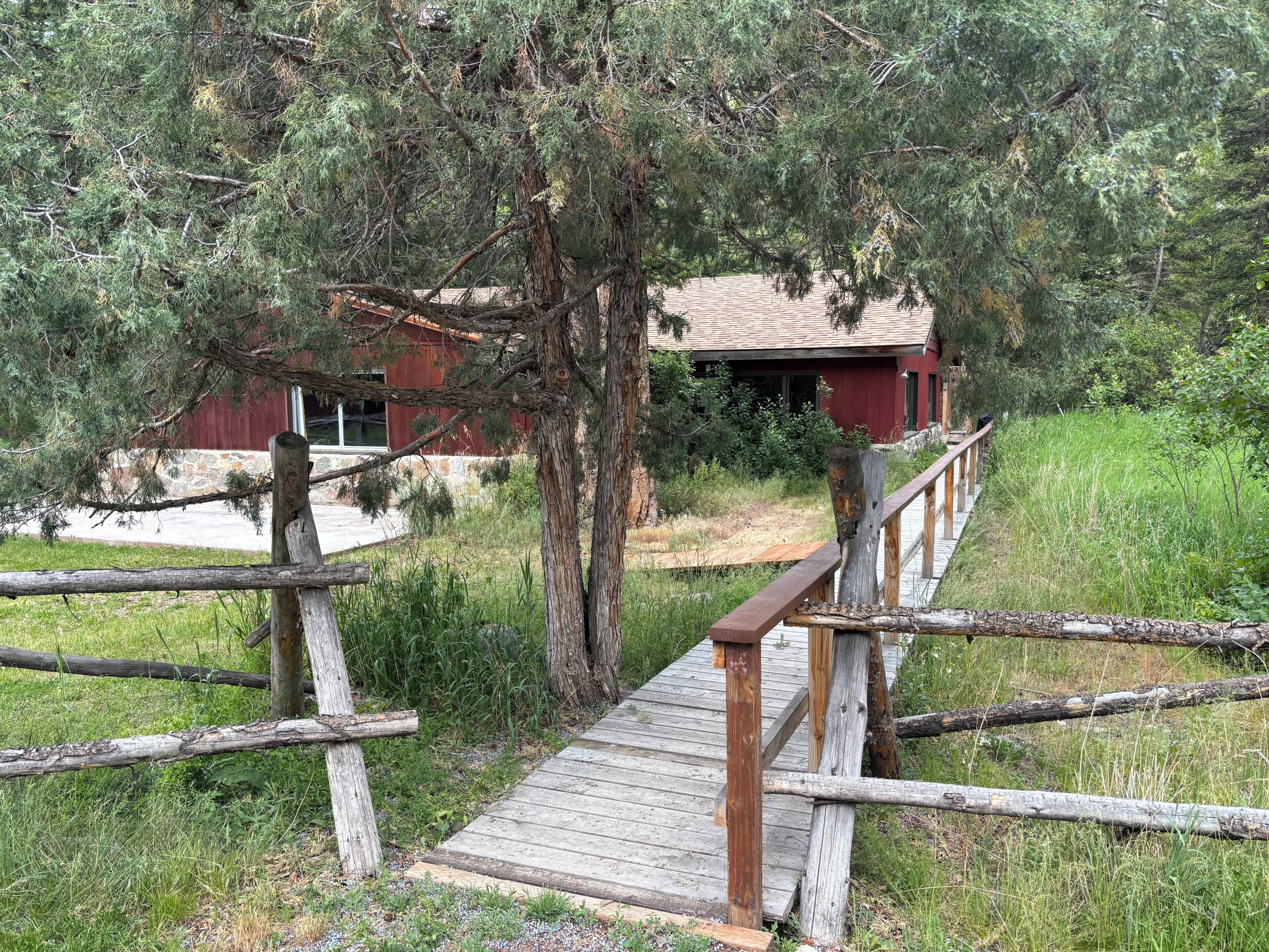 Un pont en bois mène à un bâtiment à l’extérieur rouge, entouré d’arbres et d’herbe.