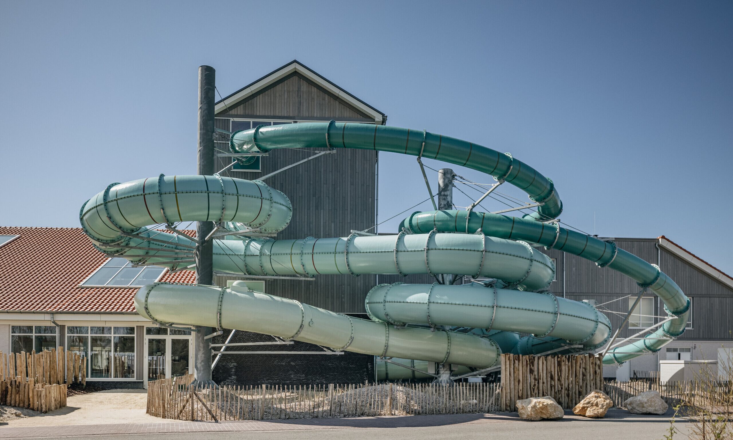 Une grande structure de toboggan aquatique se trouve devant un bâtiment doté d’un toit en pente et d’une clôture en bois.