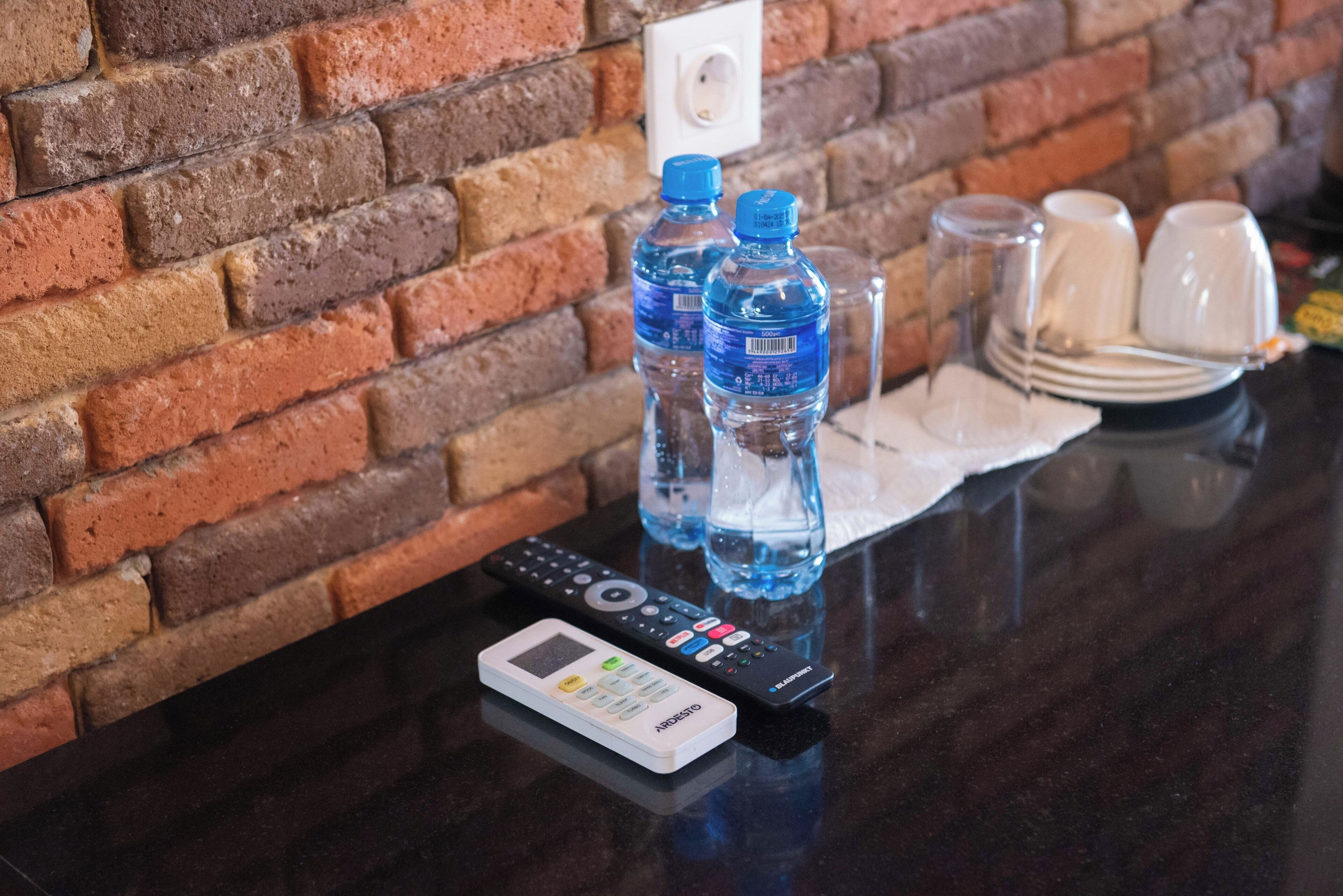 A remote control, two water bottles, and a glass on a counter with a brick wall in the background.