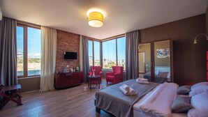 Een hotelkamer met een groot bed, een bureau met stoel, een televisie en uitzicht op de stad door grote ramen.