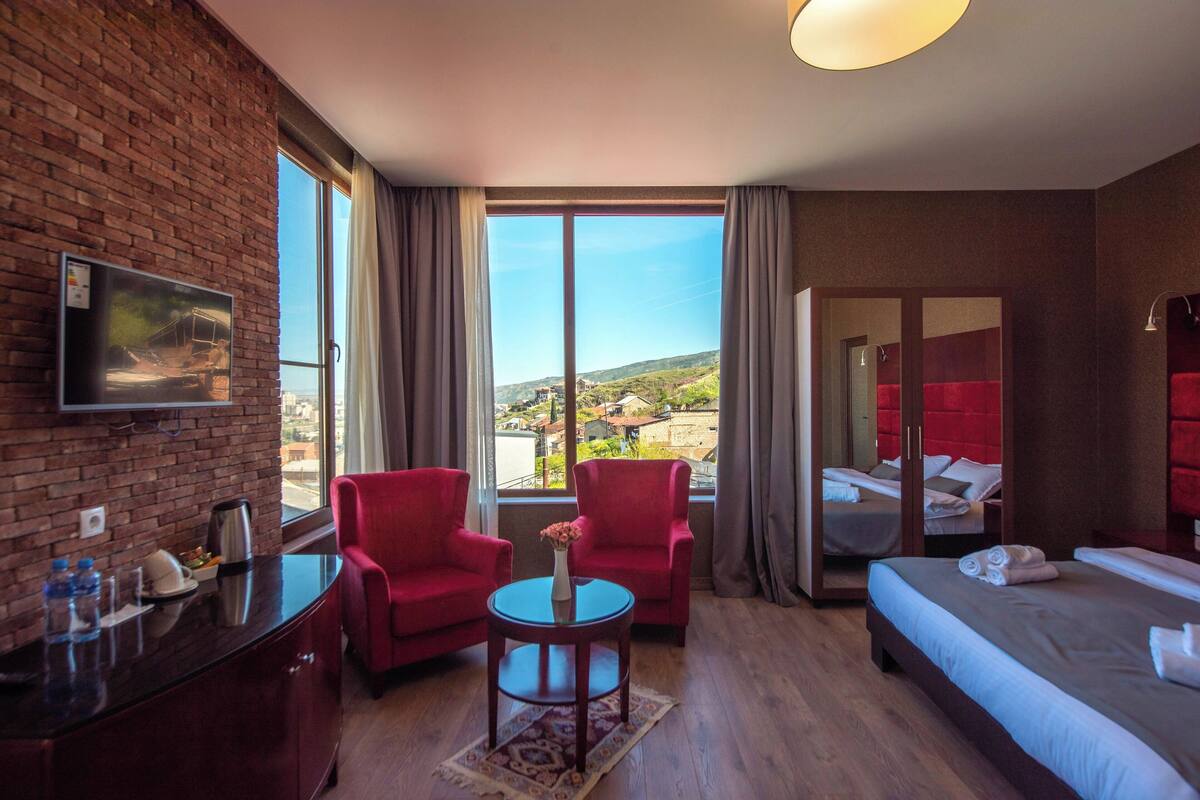 A hotel room with a brick wall, a TV, a desk with a coffee pot, two red chairs, a small table with a vase, a bed with a red headboard, and a view of a cityscape through large windows.
