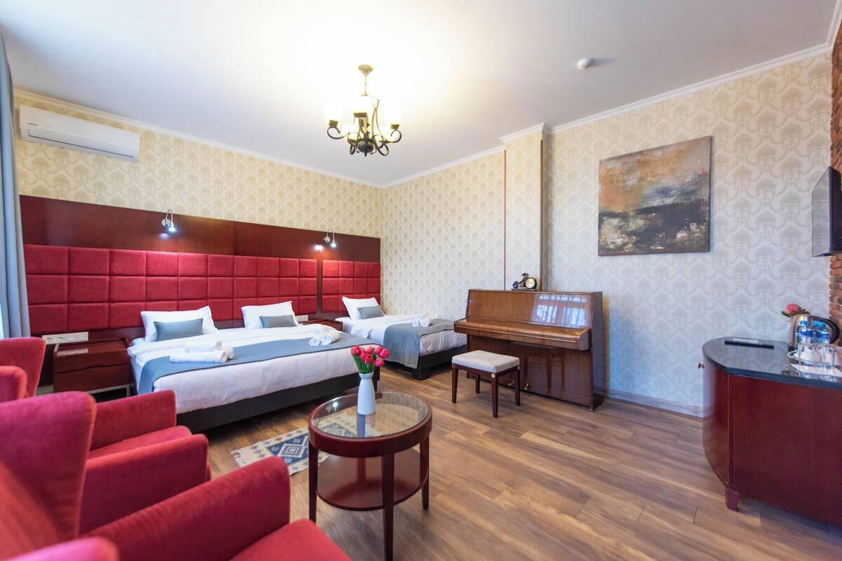 A hotel room with a large bed, a smaller bed, a piano, a red sofa, a coffee table, and a painting on the wall.