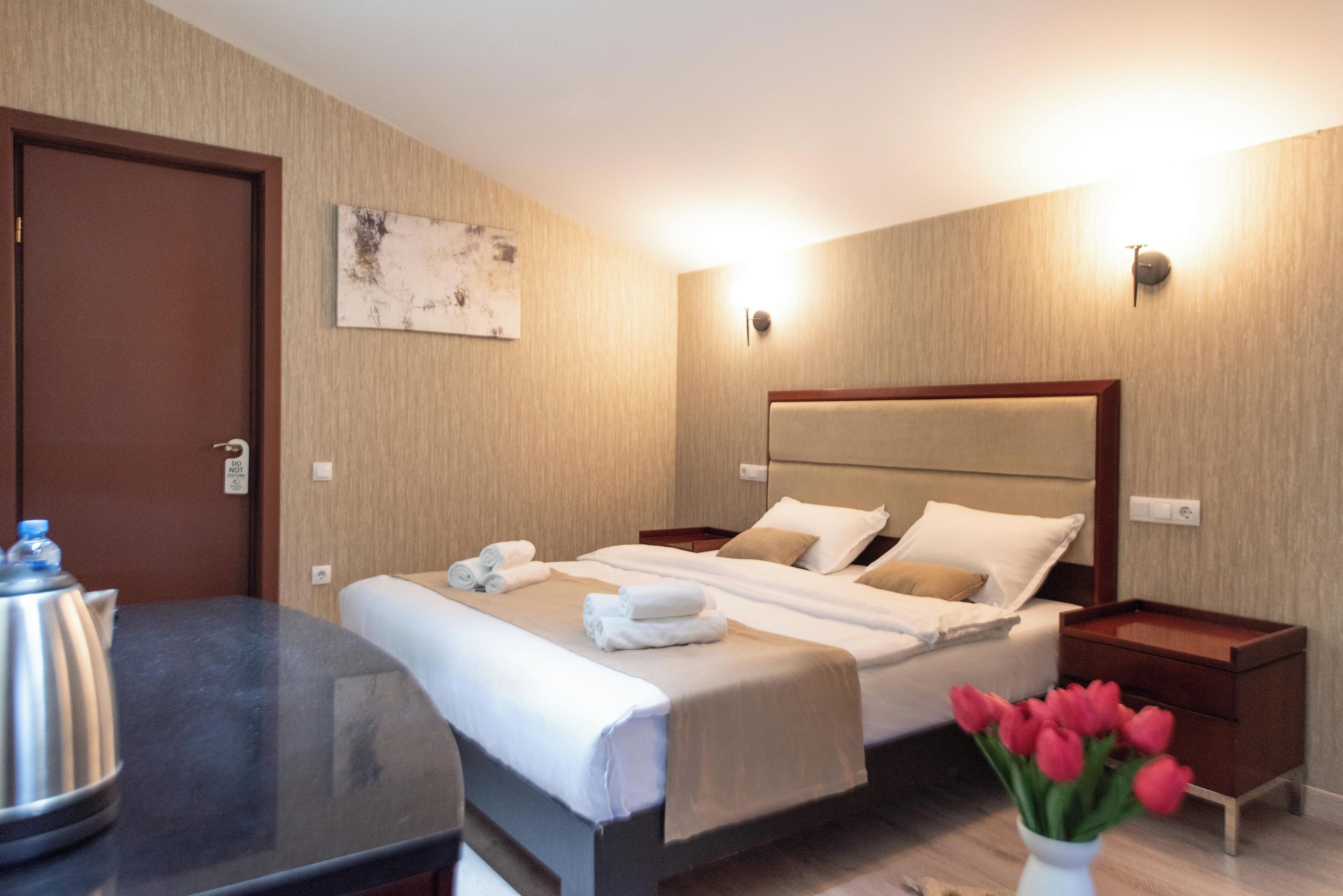 A hotel room with two beds, a bedside table with a kettle, a nightstand, and a wall-mounted light fixture.