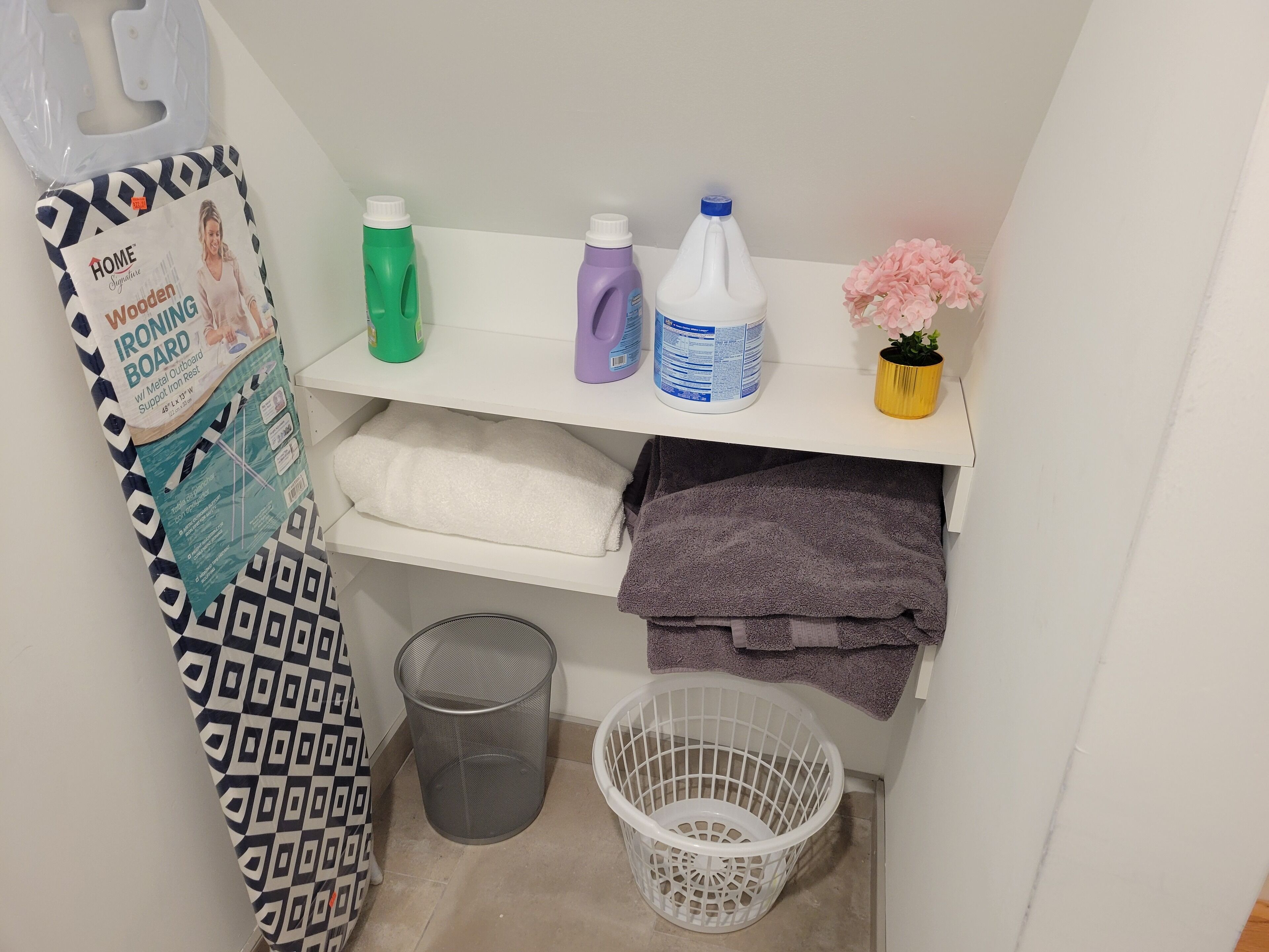 A laundry area with a metal ironing board, detergent bottles, a white laundry basket, and a trash can.
