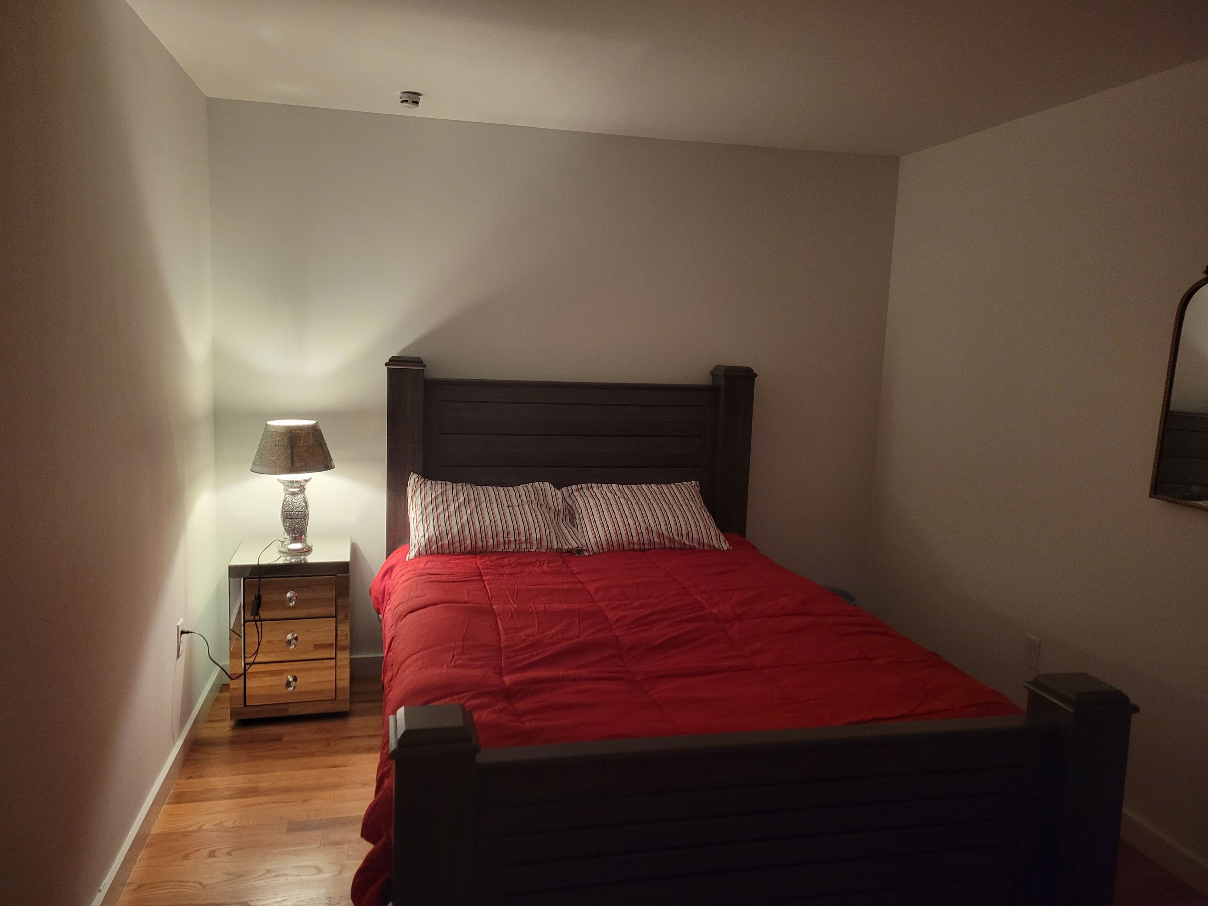 A bedroom with a wooden bed, a nightstand with a lamp, and a wall-mounted mirror.