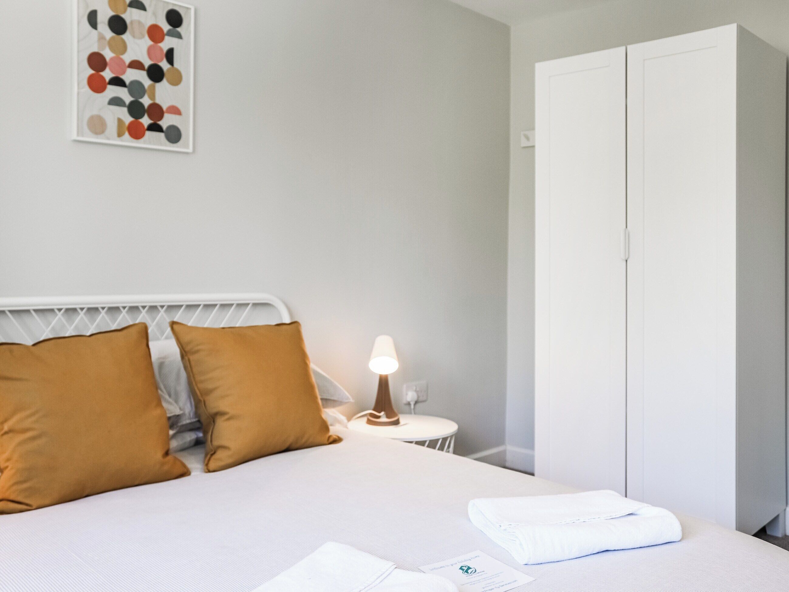 A neatly made bed with two pillows, a bedside table with a lamp, and a wardrobe.