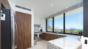A modern kitchen with wooden cabinets, a stainless steel refrigerator, a granite countertop, and a large window offering a view of the ocean.