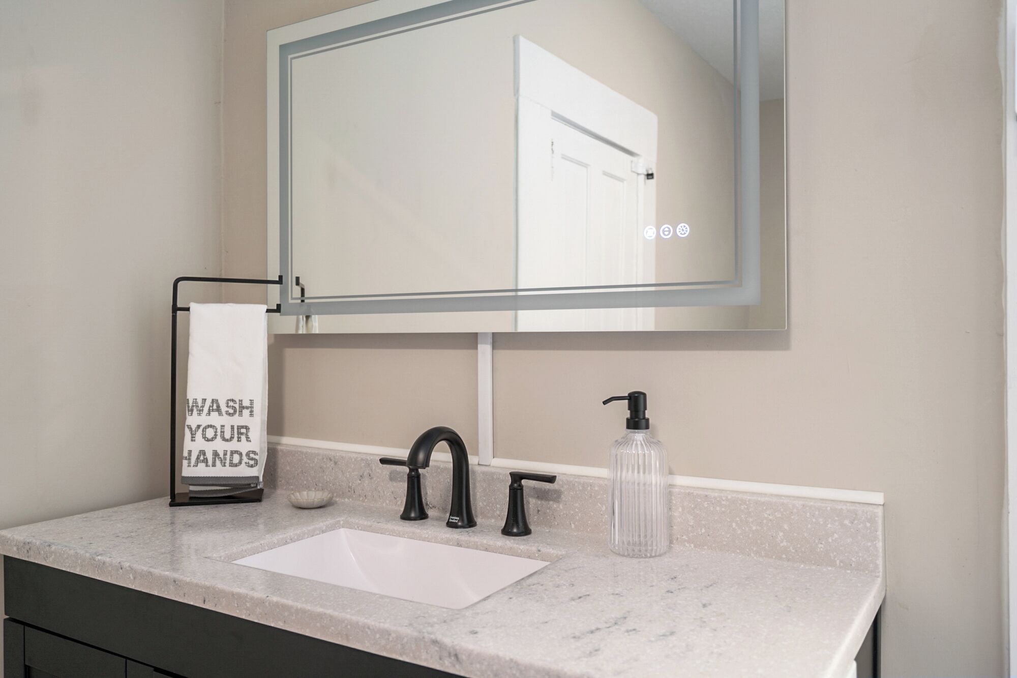 Une salle de bain avec un lavabo blanc, un robinet noir et un distributeur de savon. On peut voir un porte-serviettes sur lequel est affiché un panneau indiquant : « LAVEZ-VOUS LES MAINS ».