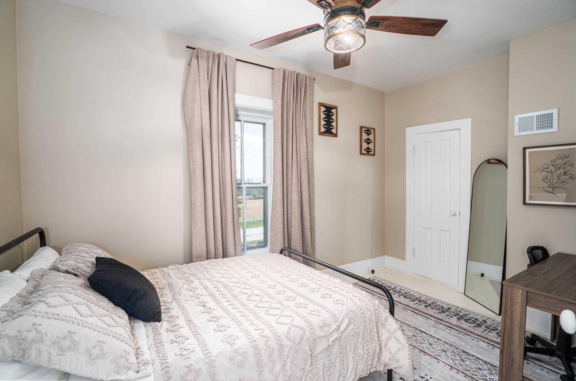 A bedroom with a bed, a ceiling fan, a window with curtains, a door, and a mirror.