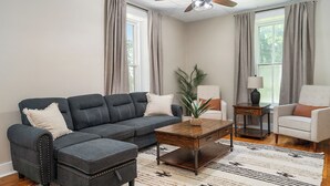 Ein Wohnzimmer mit einem Ecksofa, einem Couchtisch, einem Beistelltisch mit Lampe und einem Sessel.