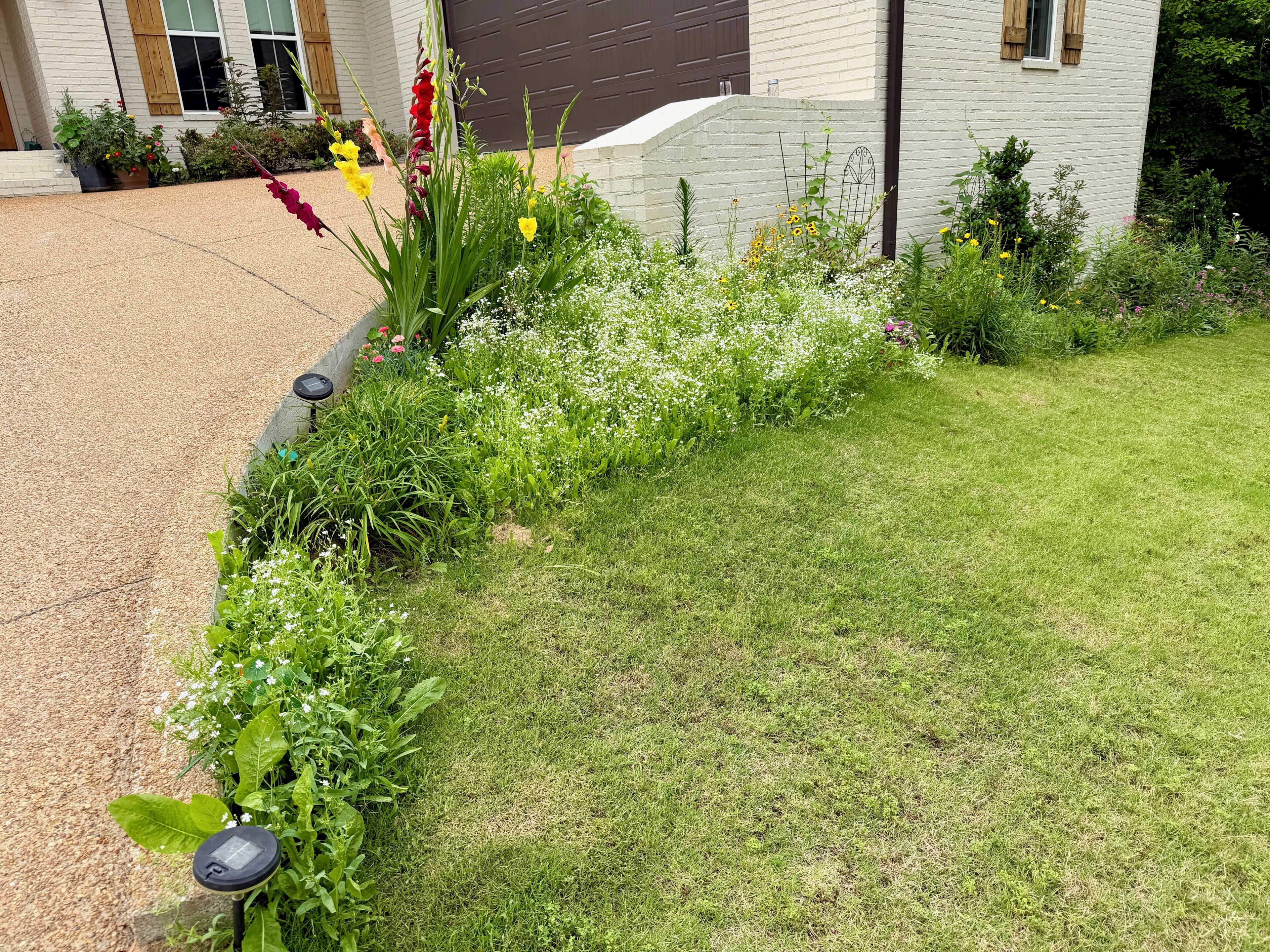 Un jardin où l’on trouve une variété de plantes et de fleurs, une pelouse et un bâtiment à la façade en briques et aux fenêtres.