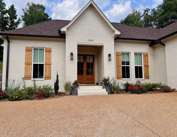 A two-story house with a white exterior, numbered 2002, featuring a front entrance with a wooden door, two windows with shutters, and a gravel driveway.