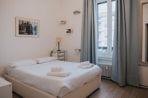 A neatly made bed with white linens and towels, a bedside table with a lamp, a framed picture on the wall, and a window with curtains.