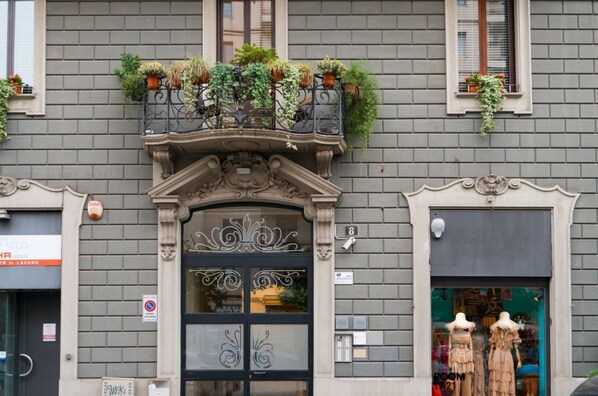 A building facade with a decorative entrance, potted plants on the balcony, and a shop window with mannequins.
