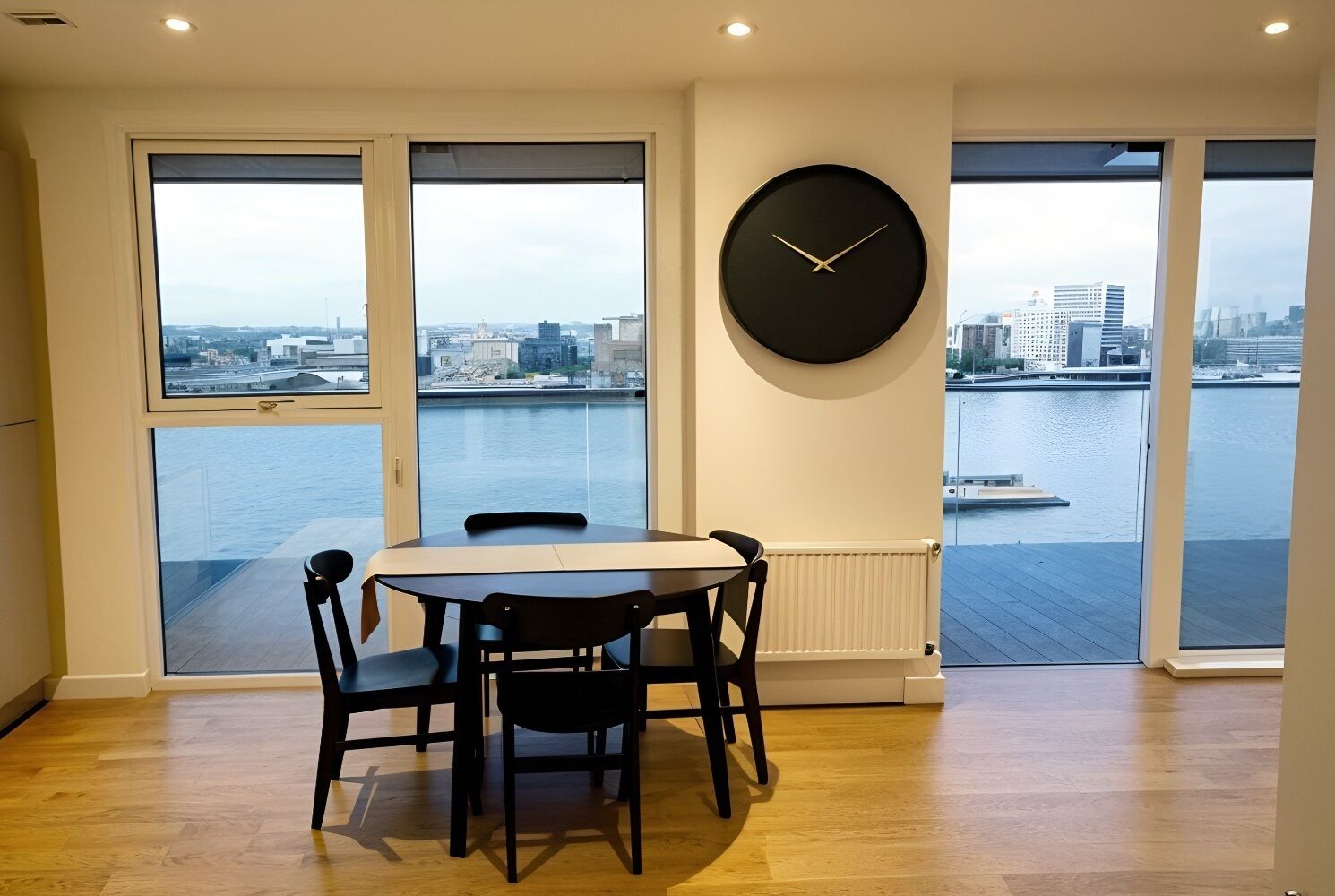 A dining area with a table and chairs, a large wall clock, and a view of a body of water with buildings in the distance.
