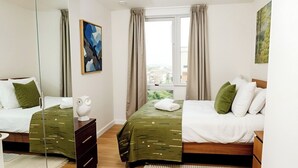 A modern bedroom with two beds, a large mirror, a window with curtains, and a view of the city.