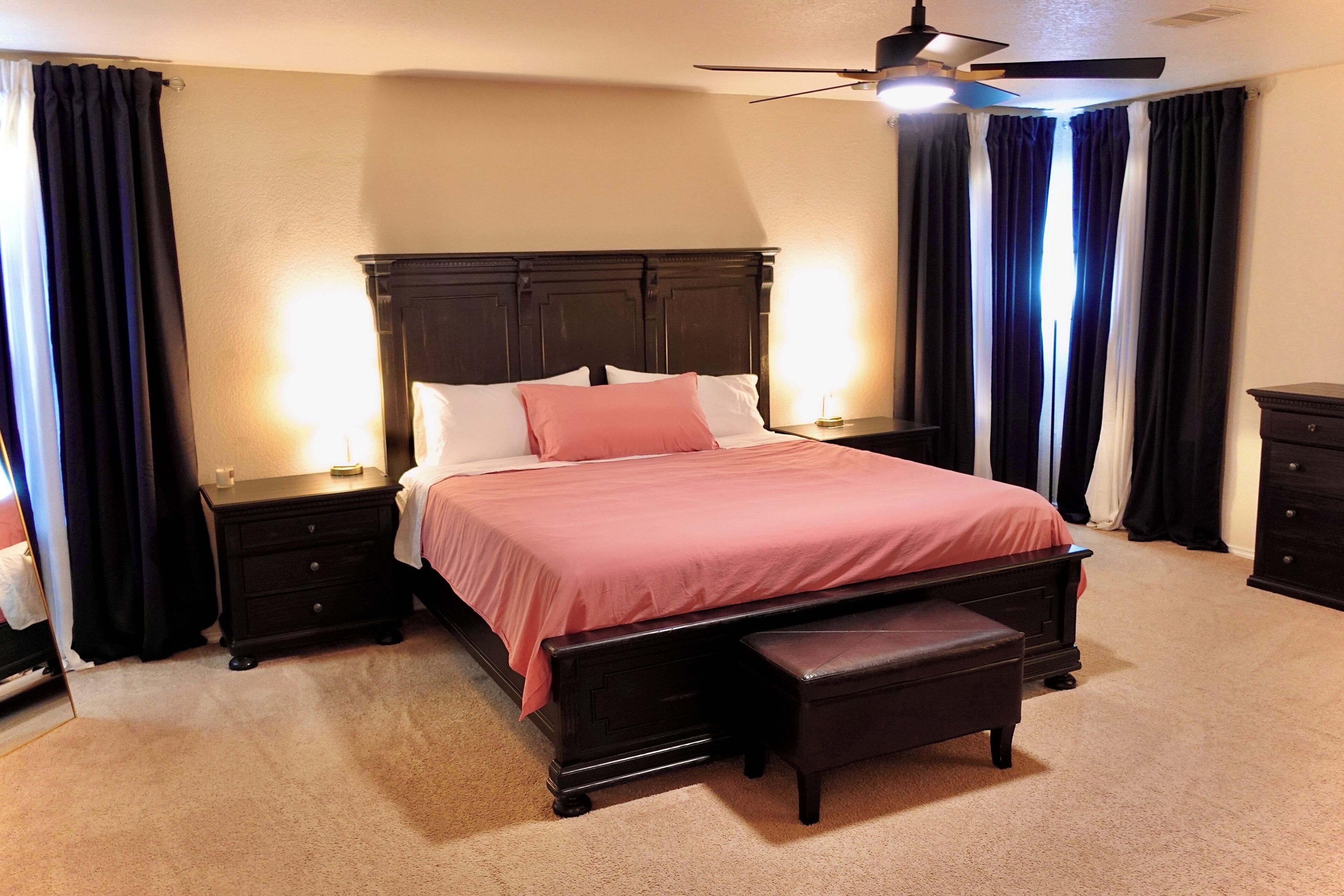 A bedroom with a large bed, a bench, a nightstand, a ceiling fan, and dark curtains.