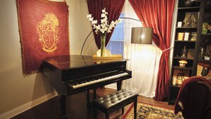 Een piano, een bank, een vaas met bloemen, een rood gordijn en een boekenkast met diverse spullen.