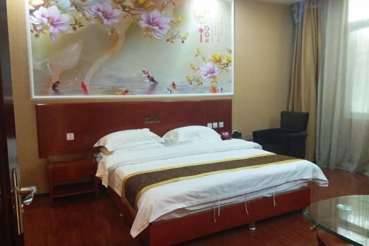 A hotel room with a bed, a wooden headboard, a glass table, and a wall with a floral painting.