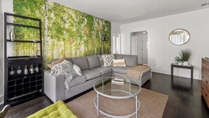 Ein Wohnzimmer mit einem grauen Ecksofa, einem Glastisch, einer schwarzen Regaleinheit mit Dekoration und einem großen Wandbild. An der Wand befindet sich ein Spiegel.