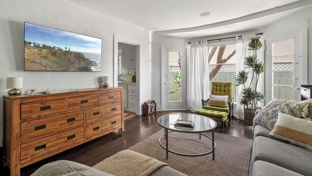 Ein Wohnzimmer mit einem Holzkabinett, einem Glastisch, einer Couch und einem Sessel. An der Wand hängt ein Gemälde, eine Pflanze steht dort, und ein Fenster ist mit Vorhängen drapiert.