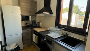 A compact kitchen with a refrigerator, microwave, sink, and a window with a view of buildings. - T2 Coloré Proche mer - Parking / Terrasse / 2 Lits 160x200 (Six-Fours-les-Plages)
