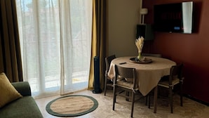 A living room with a green sofa, a dining table with chairs, a TV mounted on the wall, and a large window with curtains. - T2 Coloré Proche mer - Parking / Terrasse / 2 Lits 160x200 (Six-Fours-les-Plages)