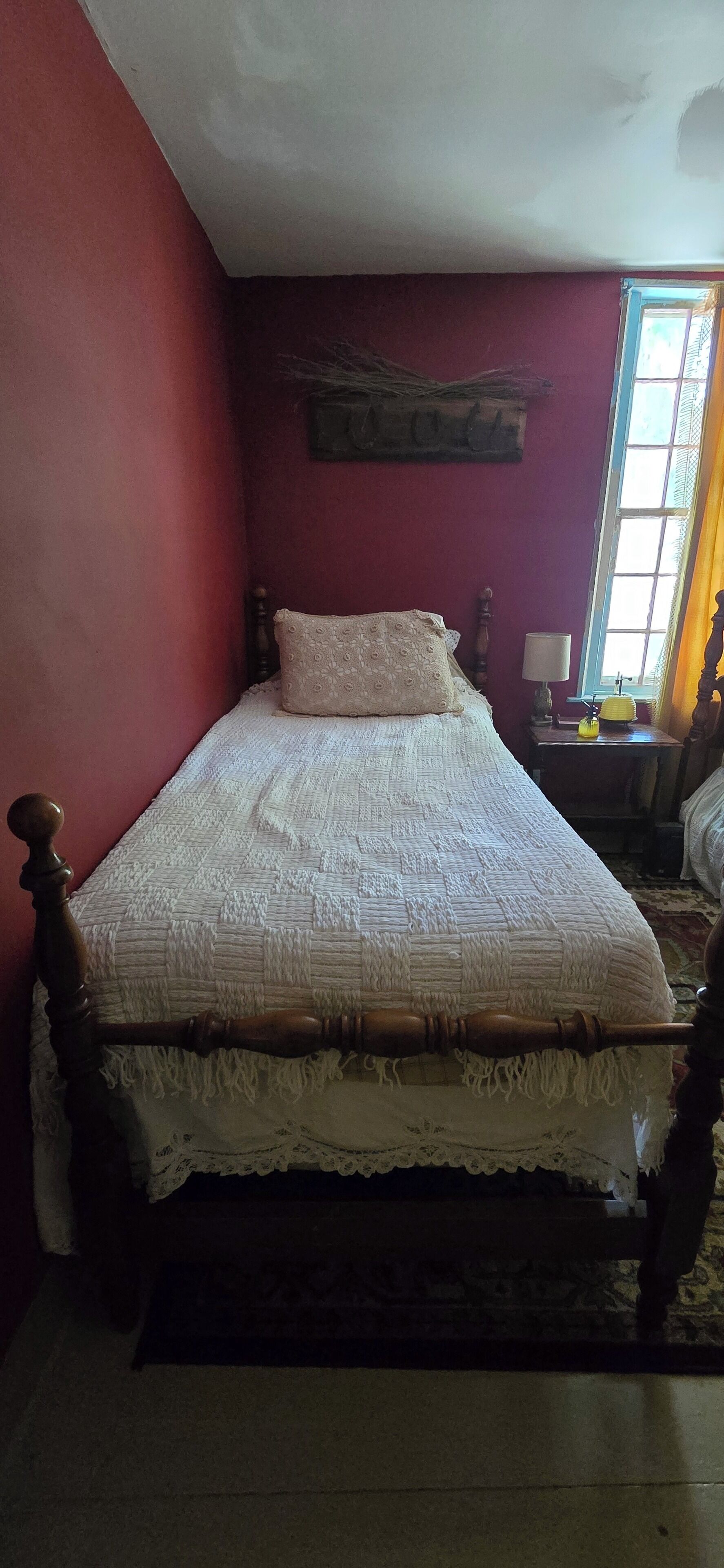Un lit simple avec un cadre en bois et une couette blanche, une table de chevet avec une lampe, et une fenêtre avec des rideaux.
