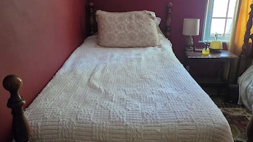 Un lit simple avec un cadre en bois et une couette blanche, une table de chevet avec une lampe, et une fenêtre avec des rideaux.