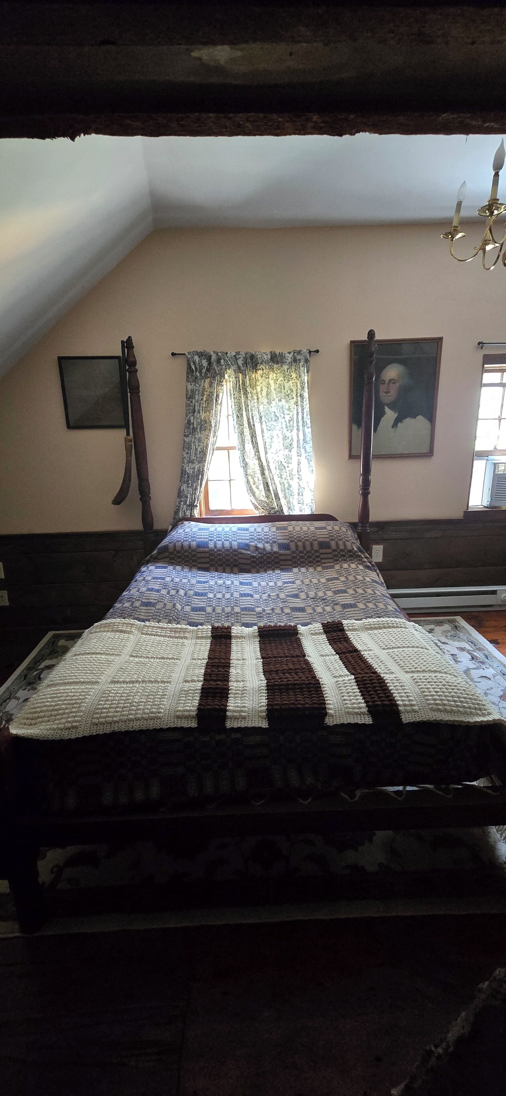 A room with a bed, a window with curtains, a painting on the wall, and a chandelier.