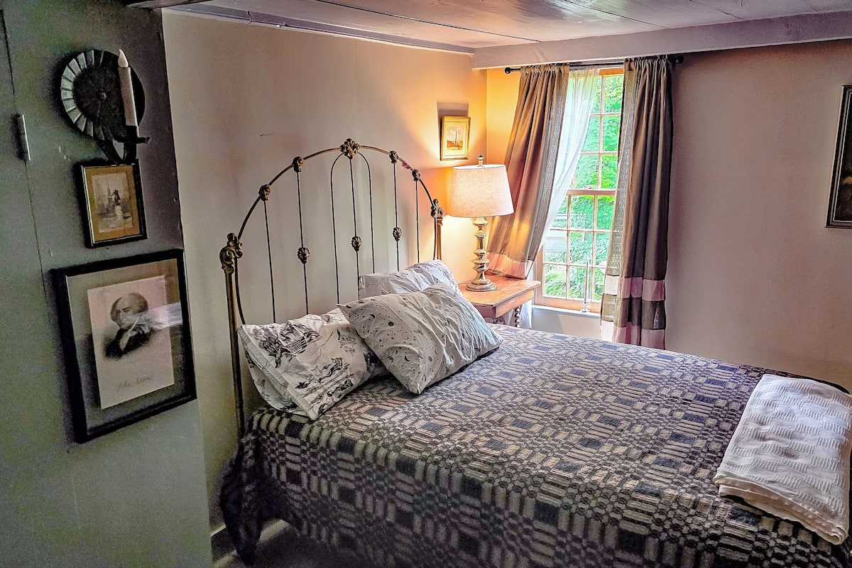 Ein Schlafzimmer mit einem Bett, einem Nachttisch mit Lampe, einem Fenster mit Vorhängen und gerahmten Bildern an den Wänden.