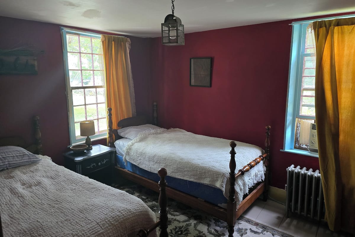 Ein Schlafzimmer mit zwei Einzelbetten, einem Holzkopfteil, einem Nachttisch, einer Lampe, einem gerahmten Bild an der Wand und einem Fenster mit Vorhängen.