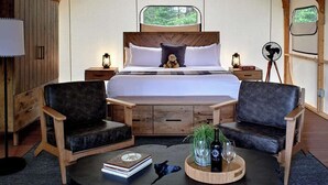 Une pièce aménagée comme une tente, avec un lit, deux chaises, une table avec des livres et une bouteille, et une petite lampe.