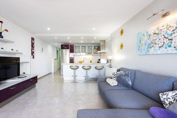 Una sala de estar moderna con un sofá gris, un televisor de pantalla plana y una cocina al fondo.